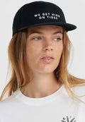 Schwarze Mütze mit flachem Schirm und gesticktem weißen Text "WIR WERDEN HIGH AUF GELOSTEN." Tragen mit einem weißen Rundhals-T-Shirt. Glatte Stoffstruktur.