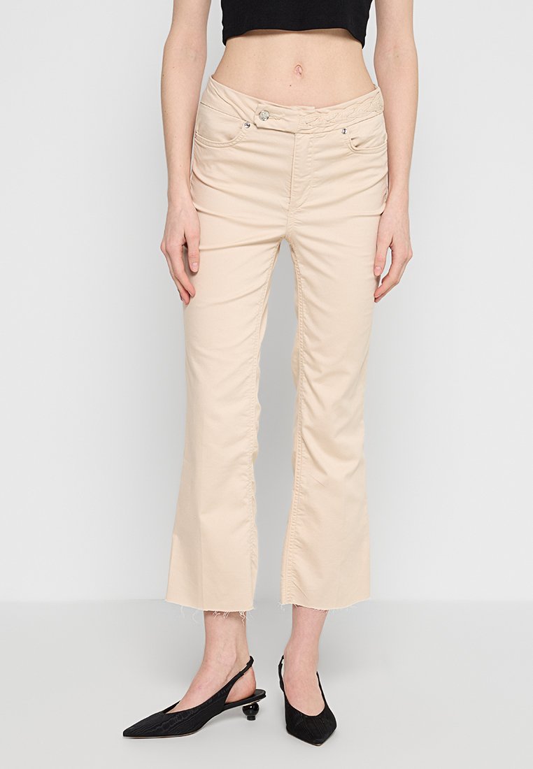 Cremefarbene, ausgestellte Hosen mit taillierter Taille, vorderen Taschen und ausgefranstem Saum. Kombiniert mit schwarzen spitzen High Heels.