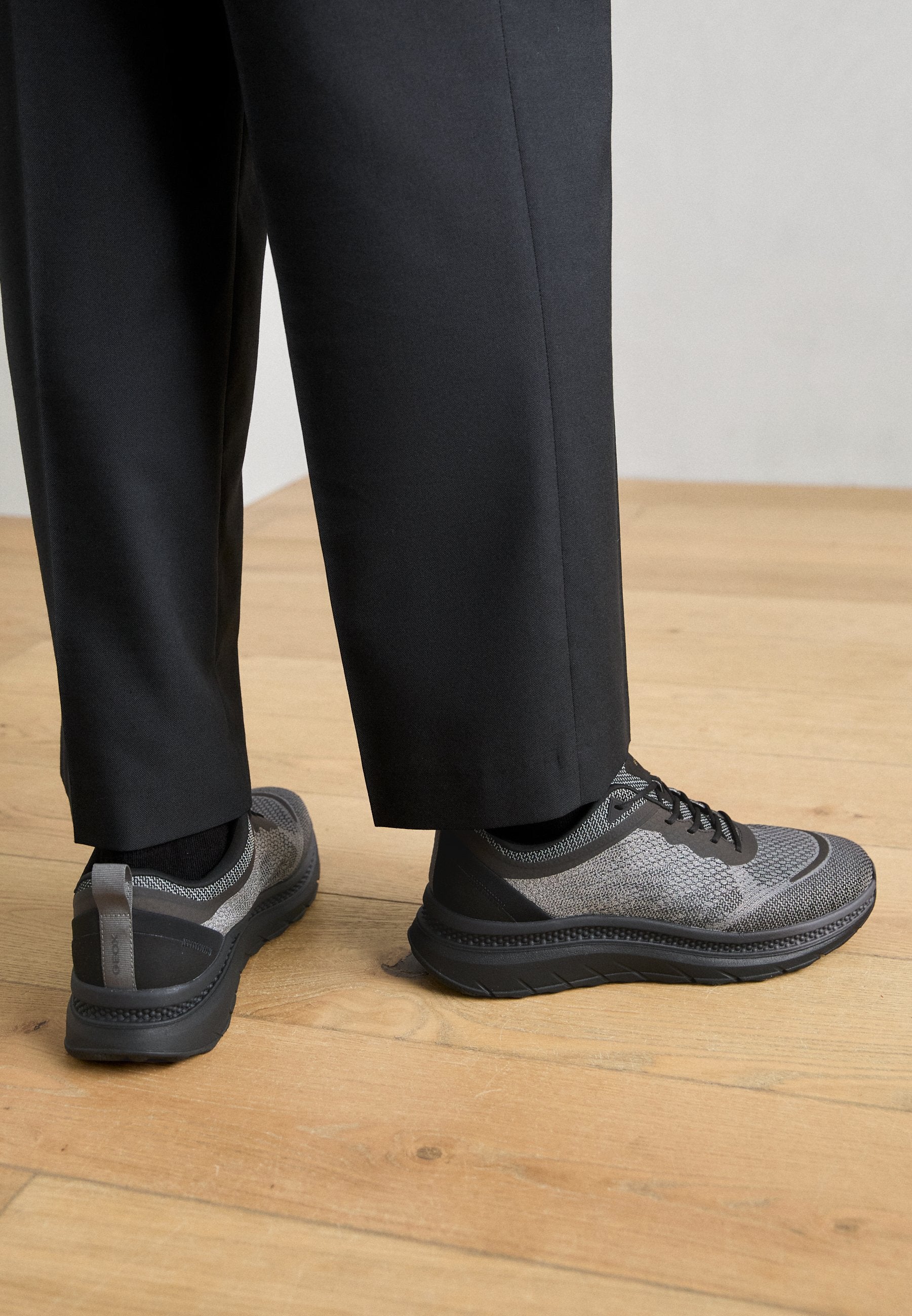 Graue Sportschuhe mit einem strukturierten Mesh-Obermaterial, schwarzen Akzenten und einer dicken Sohle, kombiniert mit maßgeschneiderten schwarzen Hosen auf einem Holzboden.