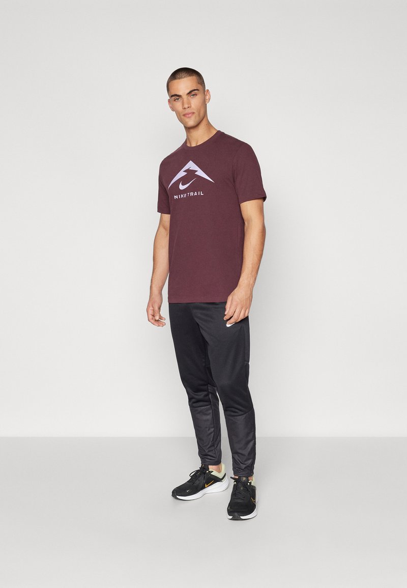 Bordeauxrotes T-Shirt mit kurzen Ärmeln und Logo, kombiniert mit schwarzen, zulaufenden Hosen und schwarzen Turnschuhen. Einfaches Design mit glatter Textur.