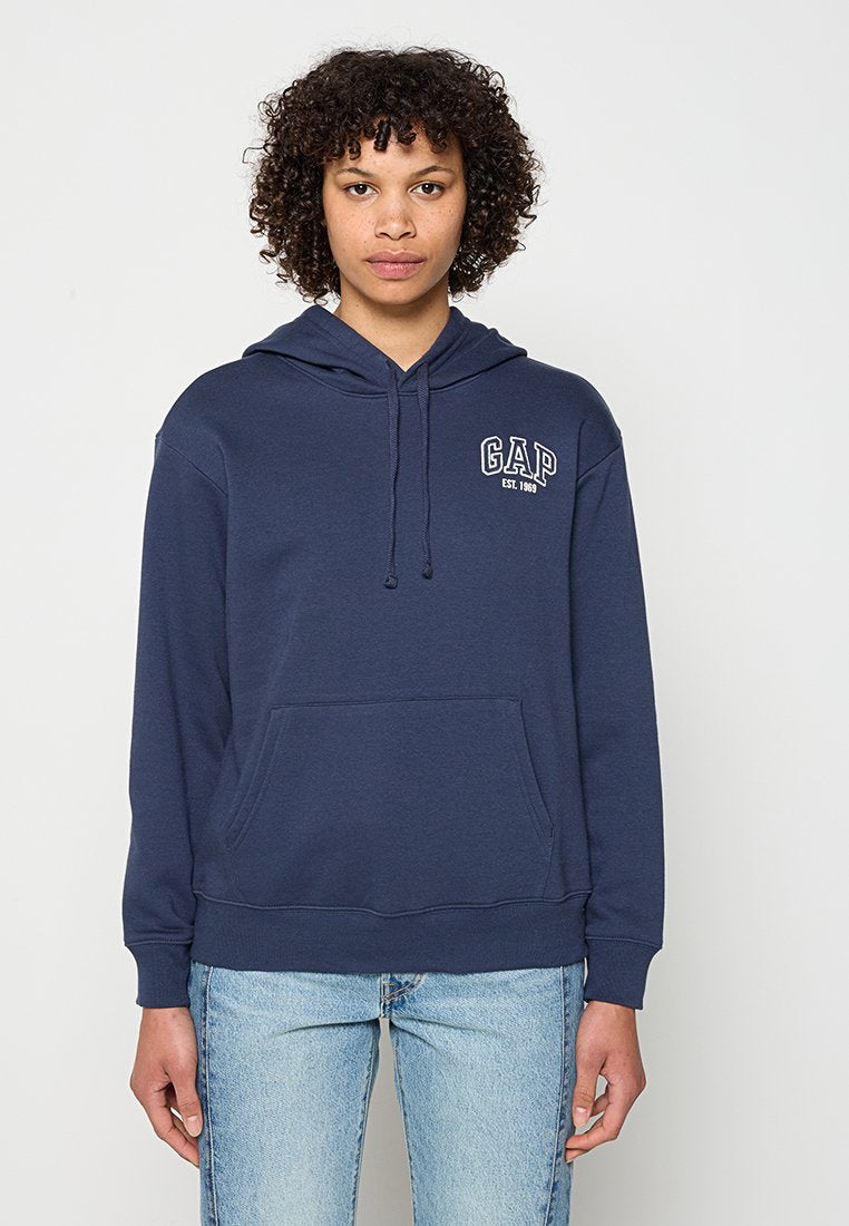 Frau mit lockigem Haar, die einen marineblauen GAP-Hoodie und hellblaue Jeans trägt und vor einem schlichten weißen Hintergrund steht.