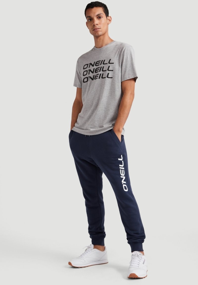 Graues T-Shirt mit schwarzem "O'NEILL"-Logo, kombiniert mit marineblauen Jogginghosen, die weißen "O'NEILL"-Text tragen. Weiße Sportschuhe runden das Outfit ab.