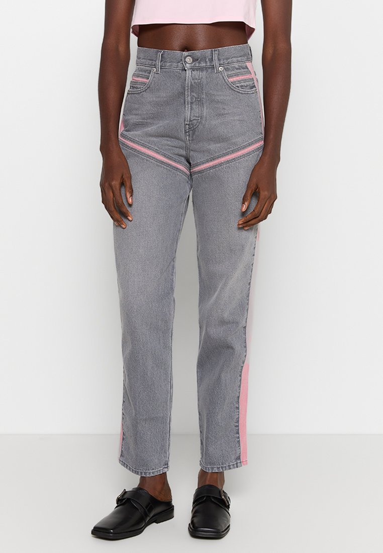 Person, der hochgeschnittene graue Jeans mit rosa Streifen an den Seiten und vorne trägt, kombiniert mit schwarzen Slippern und einem hellrosa Cropped-Top.