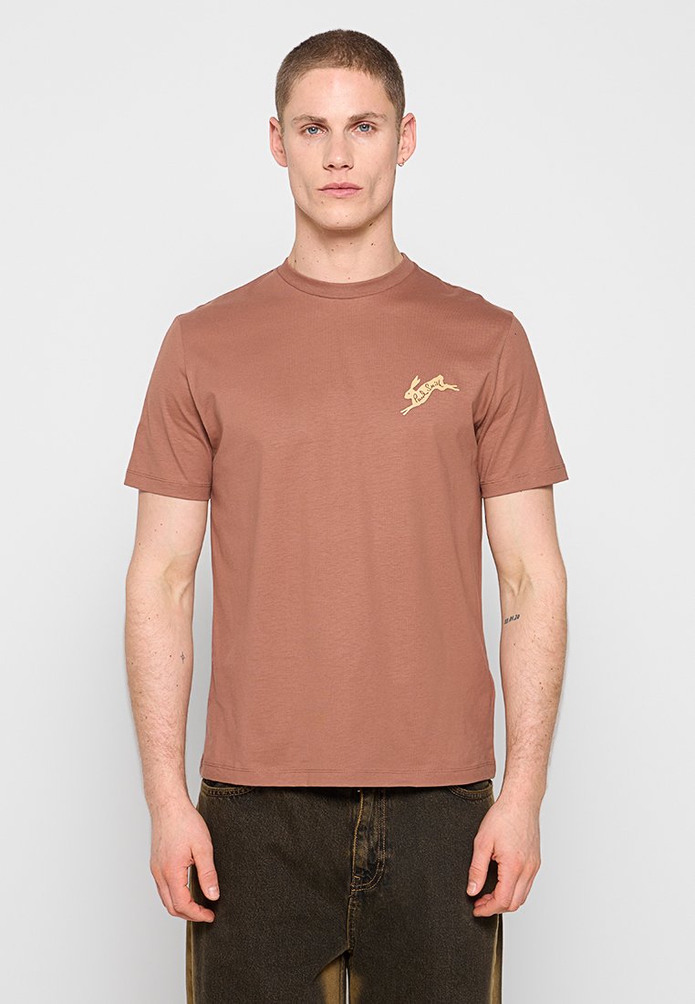 Mann trägt ein einfarbiges braunes T-Shirt mit einem kleinen gelben Hasenlogo auf der linken Brust und dunkelbraune Hosen, steht vor einem hellen Hintergrund.