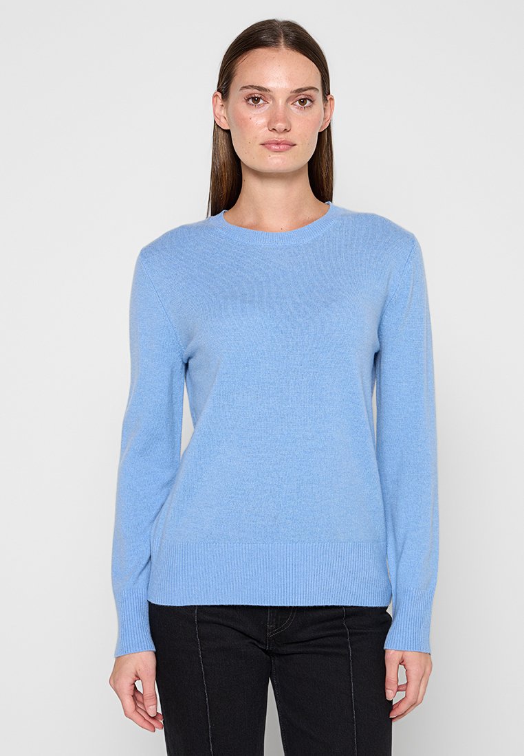 Frau mit langen braunen Haaren, die einen hellblauen Langarmpullover und schwarze Hose trägt, steht vor einem schlichten grauen Hintergrund.