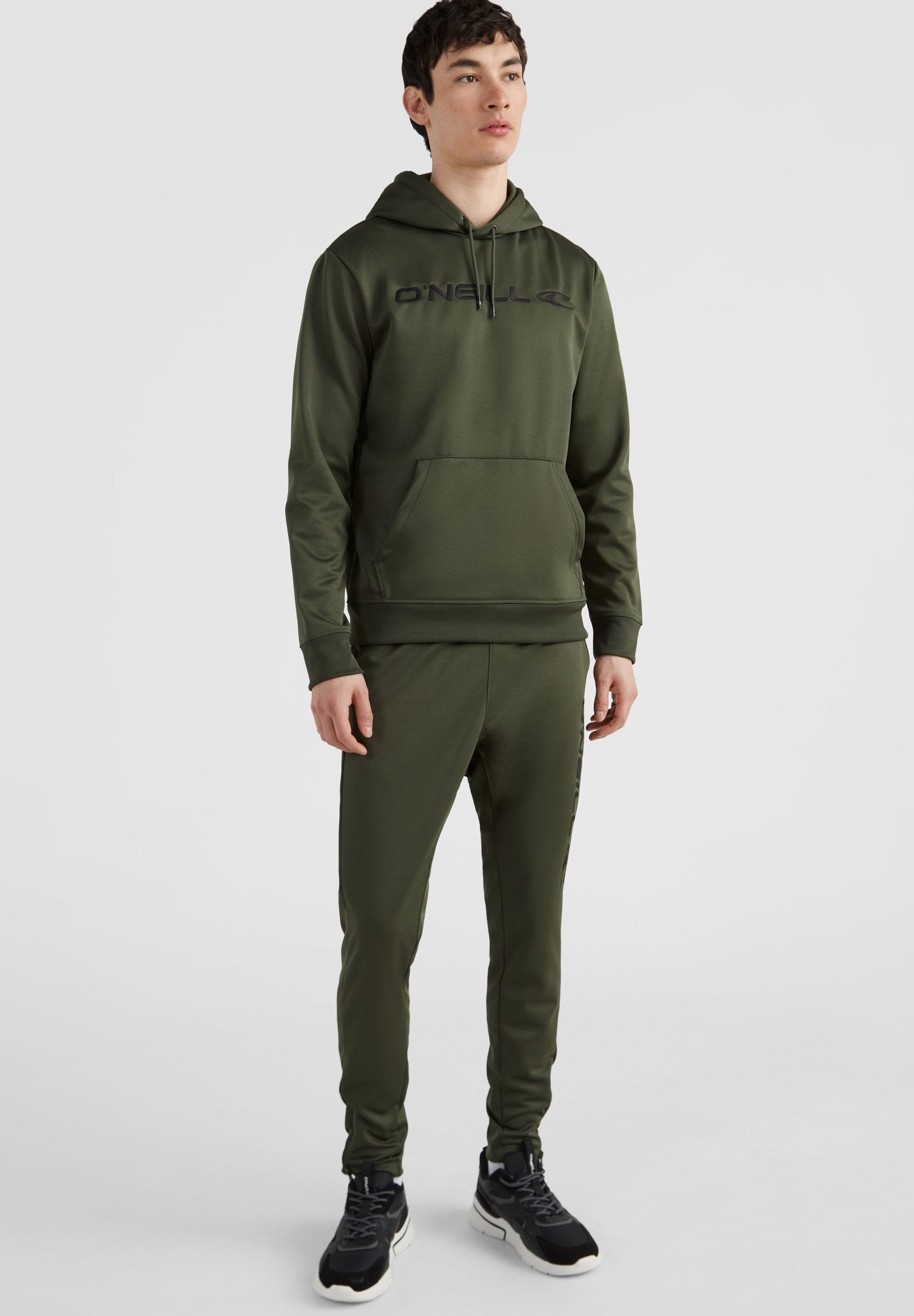 Olivgrünes Hoodie- und Jogginganzug-Set aus glattem Stoff. Mit einer Fronttasche und Logo-Text, sowie eng anliegenden Bündchen und Taillenbund. Schwarze Sneaker.