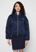 Frau mit lockigem Haar, die eine marineblaue Bomberjacke mit dem "Tommy Jeans"-Logo und einen dunklen Nadelstreifenrock trägt, steht vor schlichtem Hintergrund.