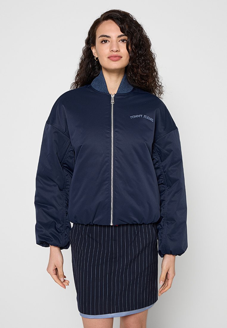 Frau mit lockigem Haar, die eine marineblaue Bomberjacke mit dem "Tommy Jeans"-Logo und einen dunklen Nadelstreifenrock trägt, steht vor schlichtem Hintergrund.