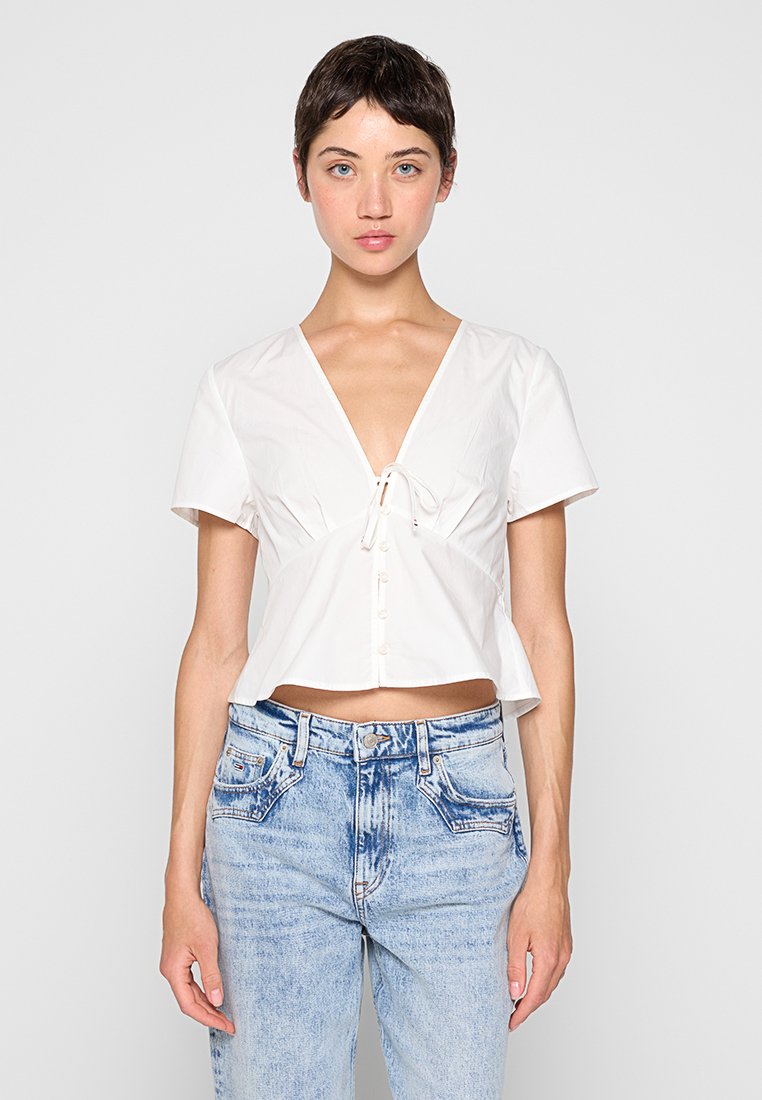 Weißes, kurzärmeliges, gecropptes Blusenshirt mit V-Ausschnitt, vorderem Bindedetail und Knopfverschluss. Kombiniert mit hellblauen, stark ausgebleichten Jeans.