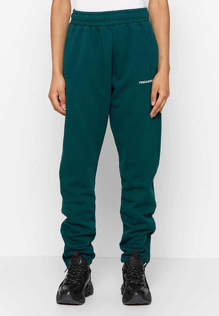 Grüne Fleece-Sweatpants mit elastischem Bund, seitlichen Taschen und einem weißen Logo auf dem linken Oberschenkel. Kombiniert mit schwarzen Sportschuhen.