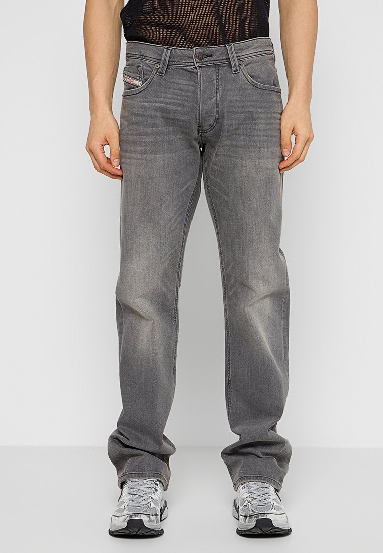 Frontalansicht einer Person in ausgewaschenen grauen Jeans, weißen und grauen Turnschuhen sowie einem schwarzen Netzoberteil, die vor einem weißen Hintergrund steht.