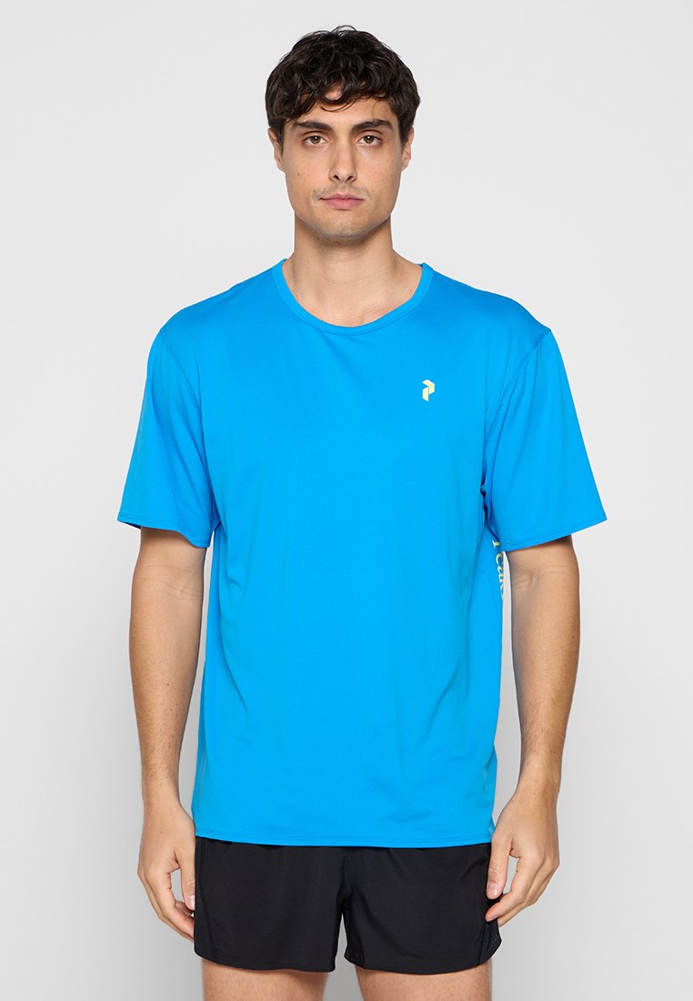 Mann trägt ein leuchtend blaues kurzärmeliges Sportshirt und schwarze Shorts und steht vor einem schlichten hellen Hintergrund.