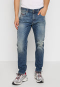 Mann trägt ausgewaschene blaue Jeans, ein weißes Trägershirt und silbergraue Laufschuhe und steht mit einer Hand in der Tasche.