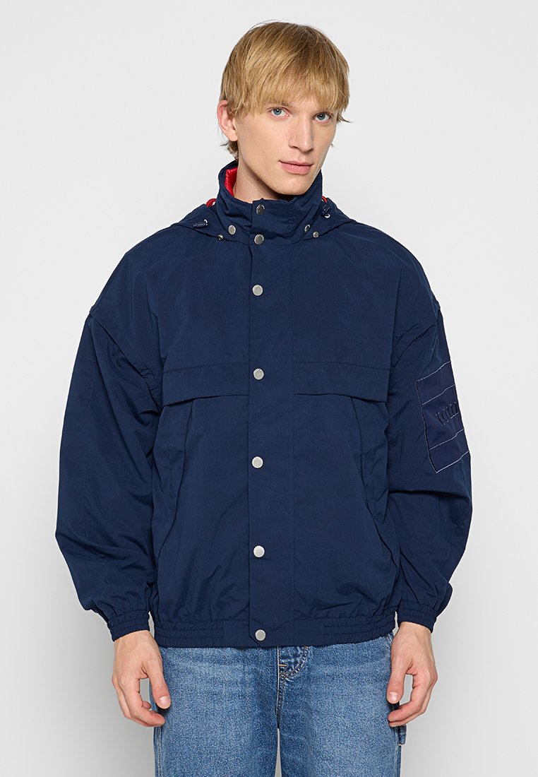Marineblaue Windjacke mit hohem Kragen, Druckknöpfen und Fronttaschen. Verfügt über elastische Bündchen und einen strukturierten Stoff mit einem Logo-Patch.