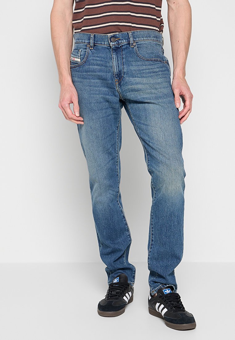 Mann in ausgewaschenen blauen Jeans, schwarzen Adidas Turnschuhen mit weißen Streifen und einem braun-beigen gestreiften Hemd, steht vor einem weißen Hintergrund.