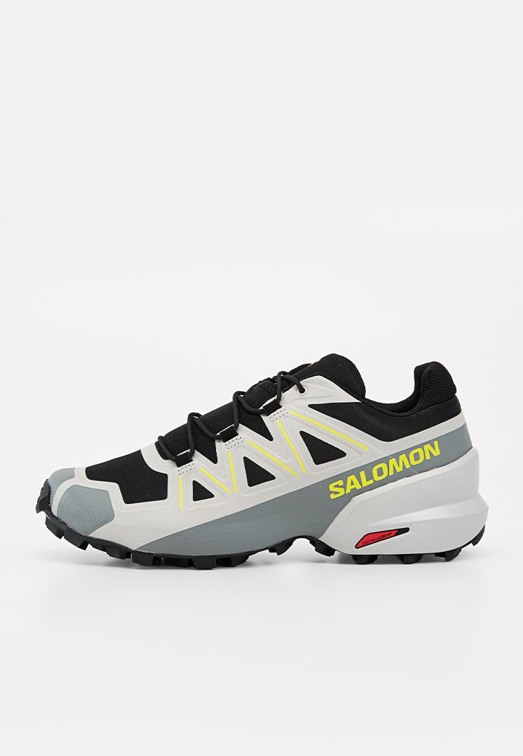 Schwarzer, grauer und weißer Salomon Trailrunning-Schuh mit gelbem Logo, gelben Akzenten und robuster schwarzer Außensohle für Grip auf unebenem Gelände.