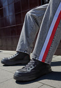 Schwarze High-Top-Sneaker mit Netzpaneelen und dicken Gummisohlen, kombiniert mit grauen Jeans mit roten und weißen Seitenstreifen.