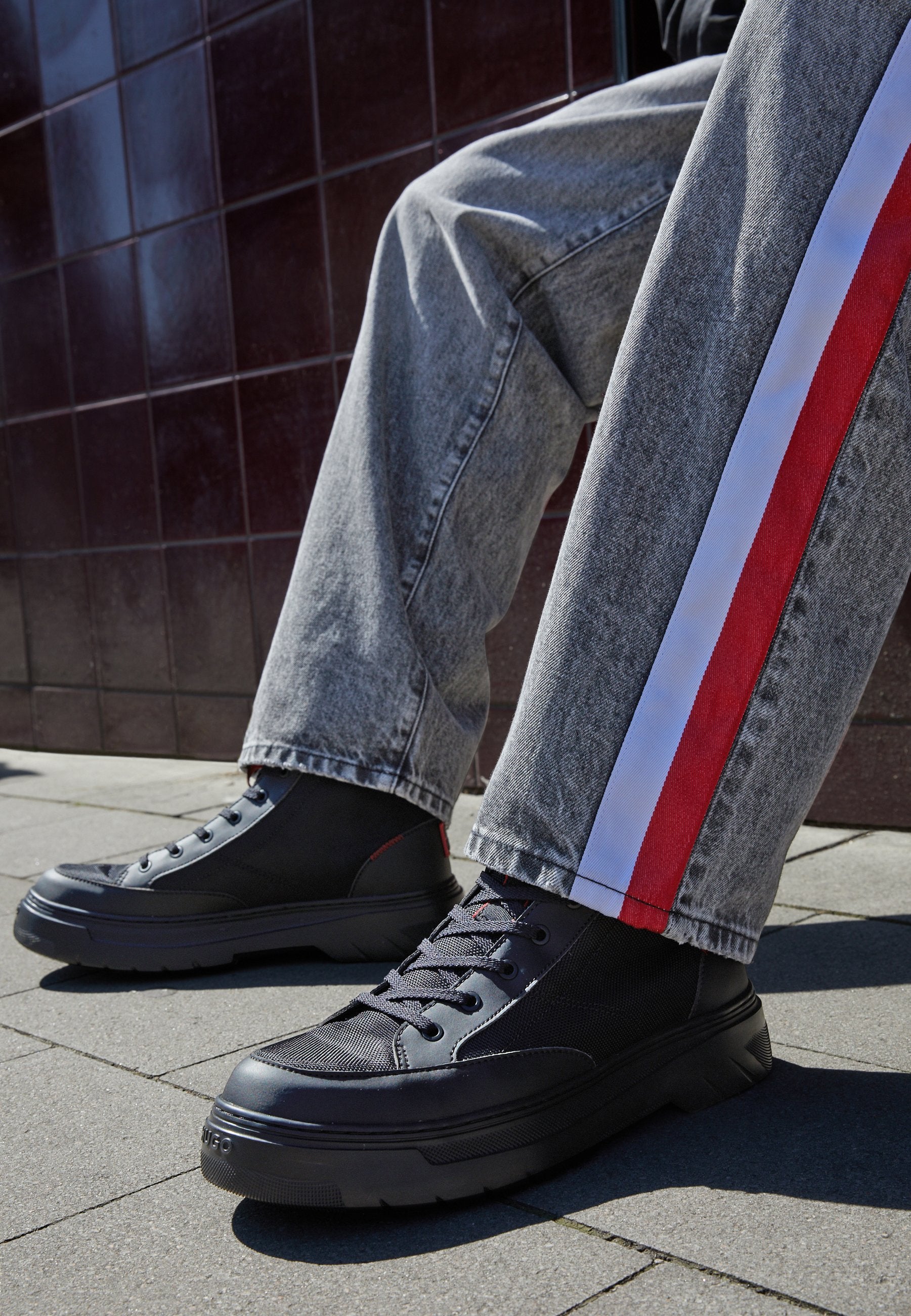 Schwarze High-Top-Sneaker mit Netzpaneelen und dicken Gummisohlen, kombiniert mit grauen Jeans mit roten und weißen Seitenstreifen.