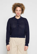 Frau mit lockigem Haar, die einen marineblauen Strickcardigan mit Knöpfen und beige Hosen trägt, steht vor einem schlichten hellen Hintergrund.