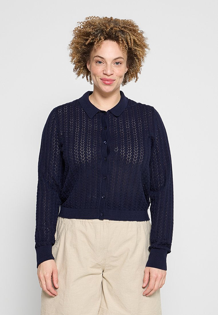 Frau mit lockigem Haar, die einen marineblauen Strickcardigan mit Knöpfen und beige Hosen trägt, steht vor einem schlichten hellen Hintergrund.