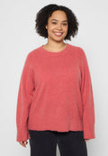 Lächelnde Frau mit lockigem Haar, die einen locker sitzenden rosa Pullover und schwarze Hosen trägt, vor einem schlichten hellen Hintergrund steht.
