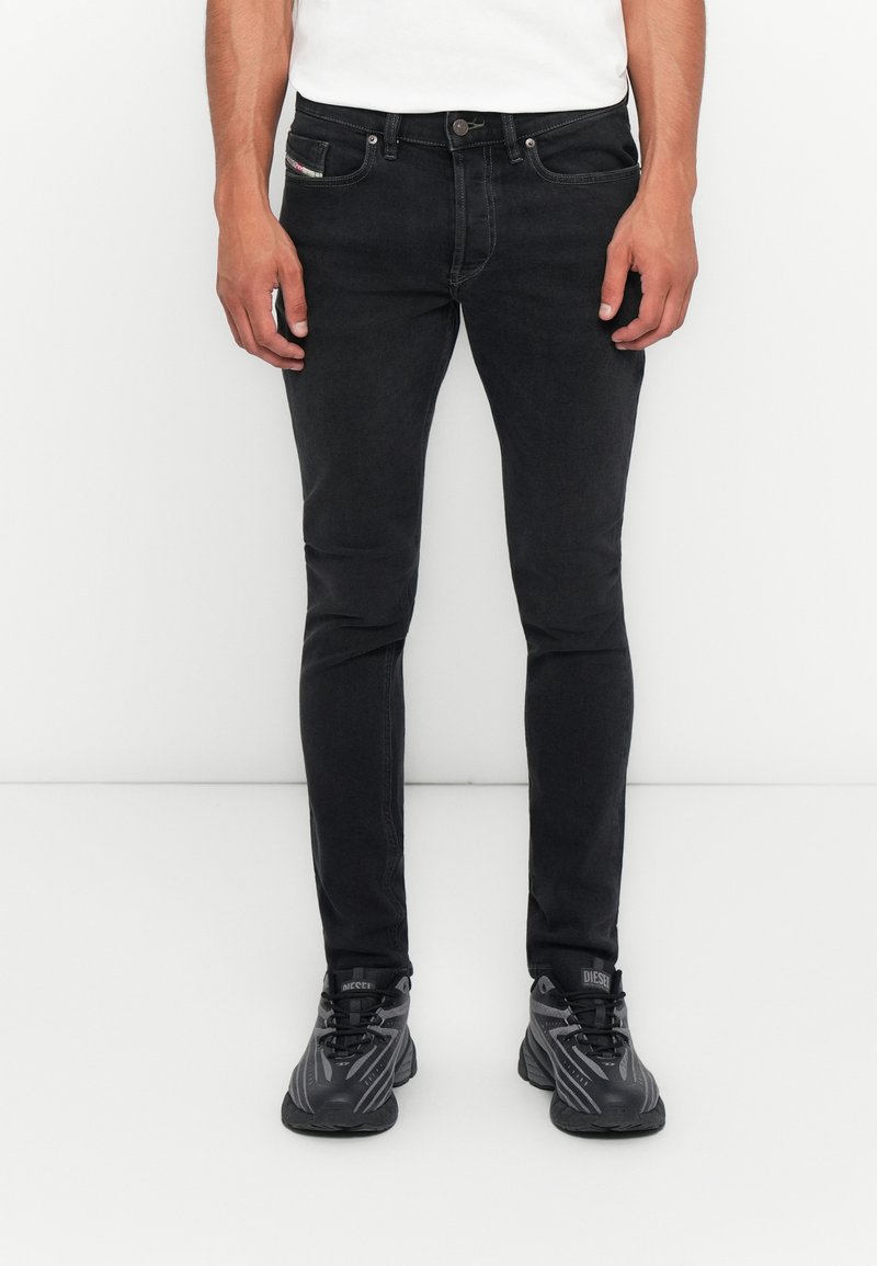 Mann in eng anliegenden schwarzen Jeans, weißem Hemd und schwarzen Diesel-Sneakern, der vor einem schlichten weißen Hintergrund steht.