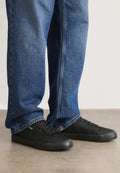 Schwarze Leinensneaker mit Gummisohlen und runder Zehenpartie. Schnürdesign mit einem kleinen weißen Logo-Detail an der Seite. Getragen mit blauen Jeans.