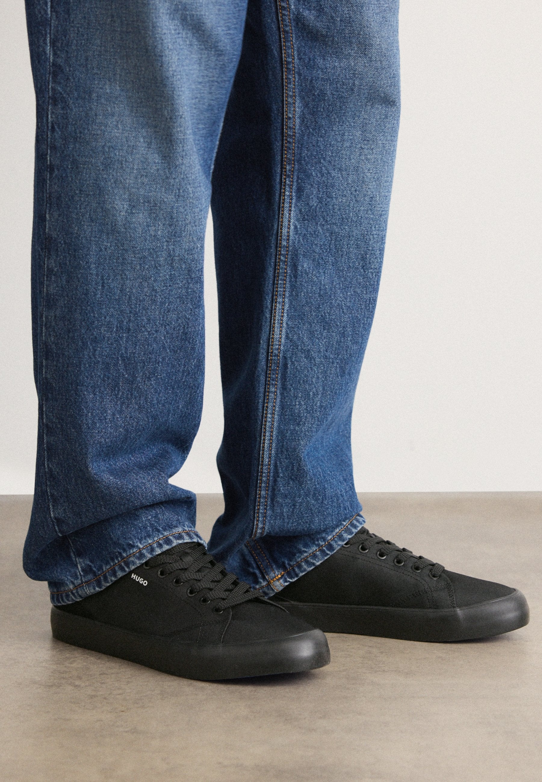 Schwarze Leinensneaker mit Gummisohlen und runder Zehenpartie. Schnürdesign mit einem kleinen weißen Logo-Detail an der Seite. Getragen mit blauen Jeans.