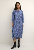 Blaues Kleid im Paisleymuster mit 3/4-Ärmeln, lockerer Passform und knielangem Saum. Kombiniert mit schwarzen Stiefeletten, vor einem neutralen Hintergrund.