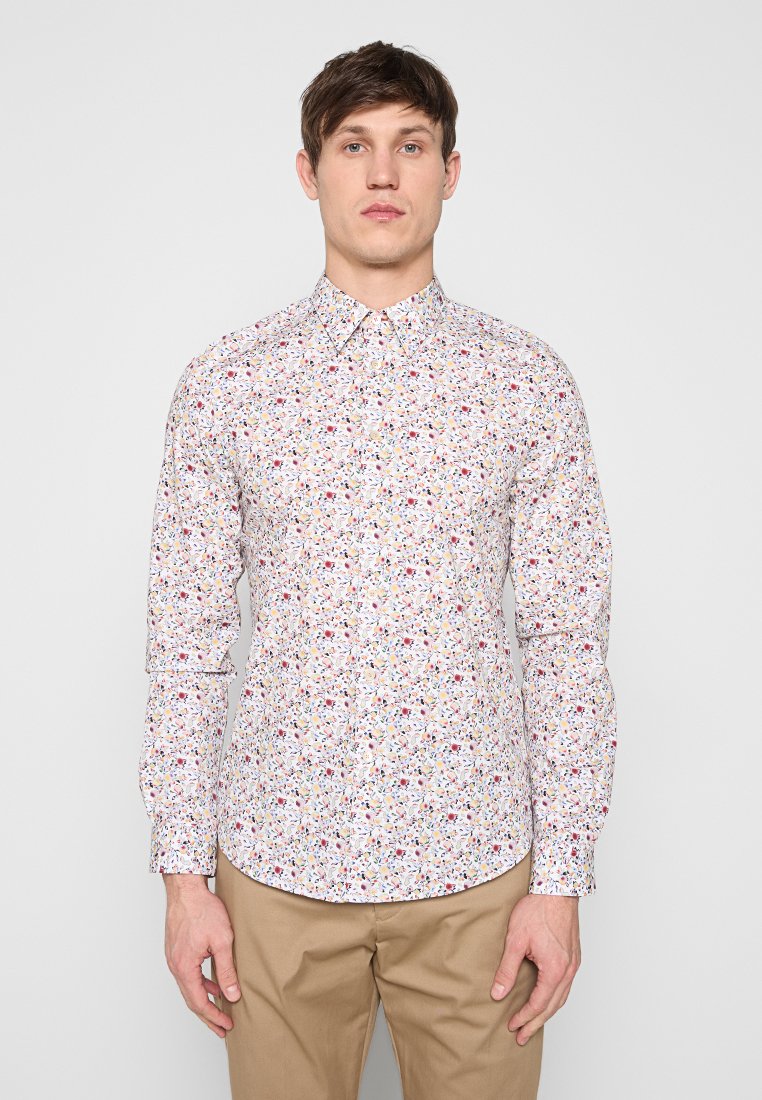 Mann trägt ein langärmliges, geknöpftes Hemd mit buntem kleinem Blumenmuster und beigen Hosen, steht vor hellem einfarbigem Hintergrund.