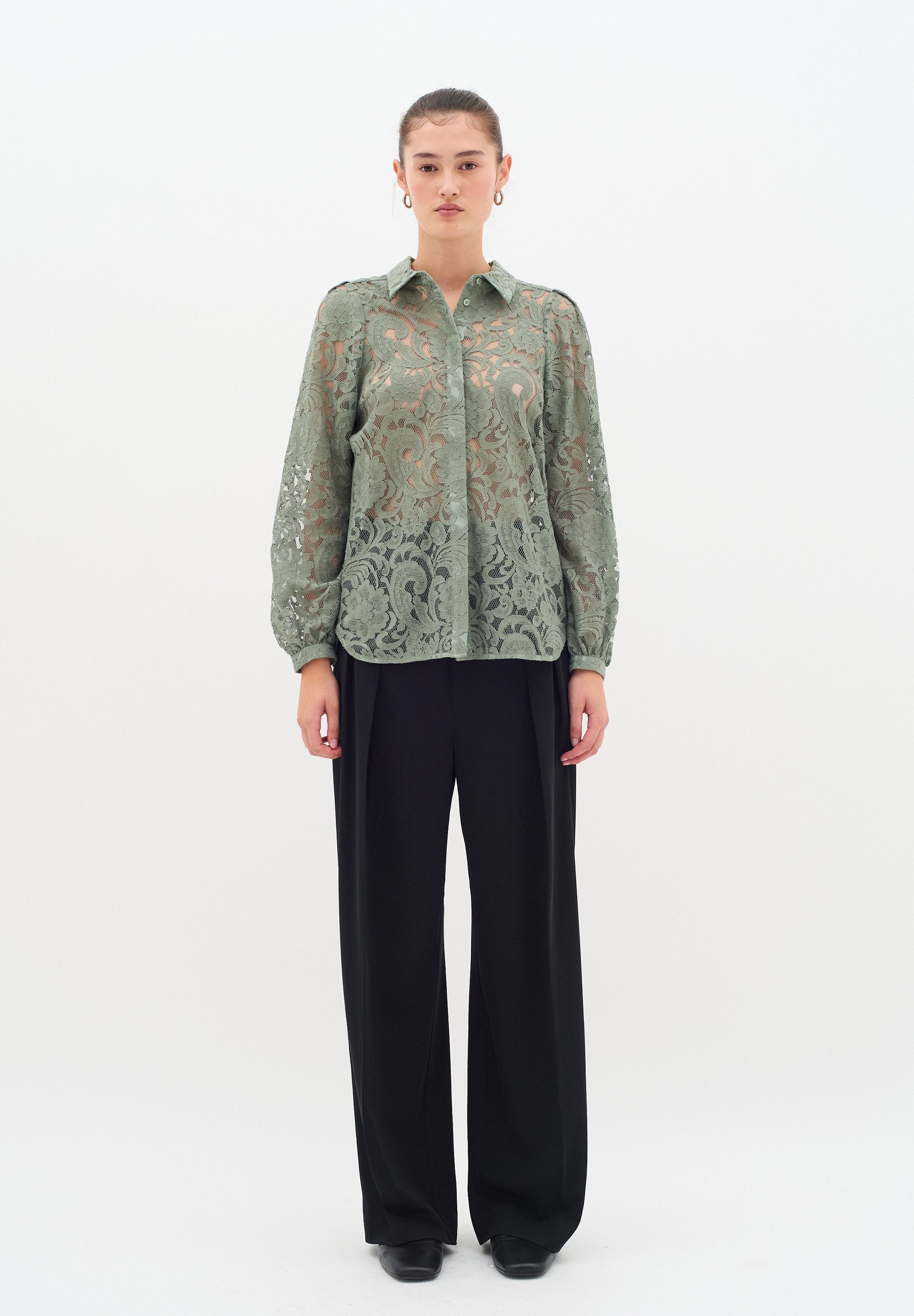 Durchsichtige grüne Spitzenbluse mit floralem Muster, Knopfverschluss, langen Ärmeln und abgerundetem Saum, kombiniert mit hochgeschnittenen schwarzen Hosen.