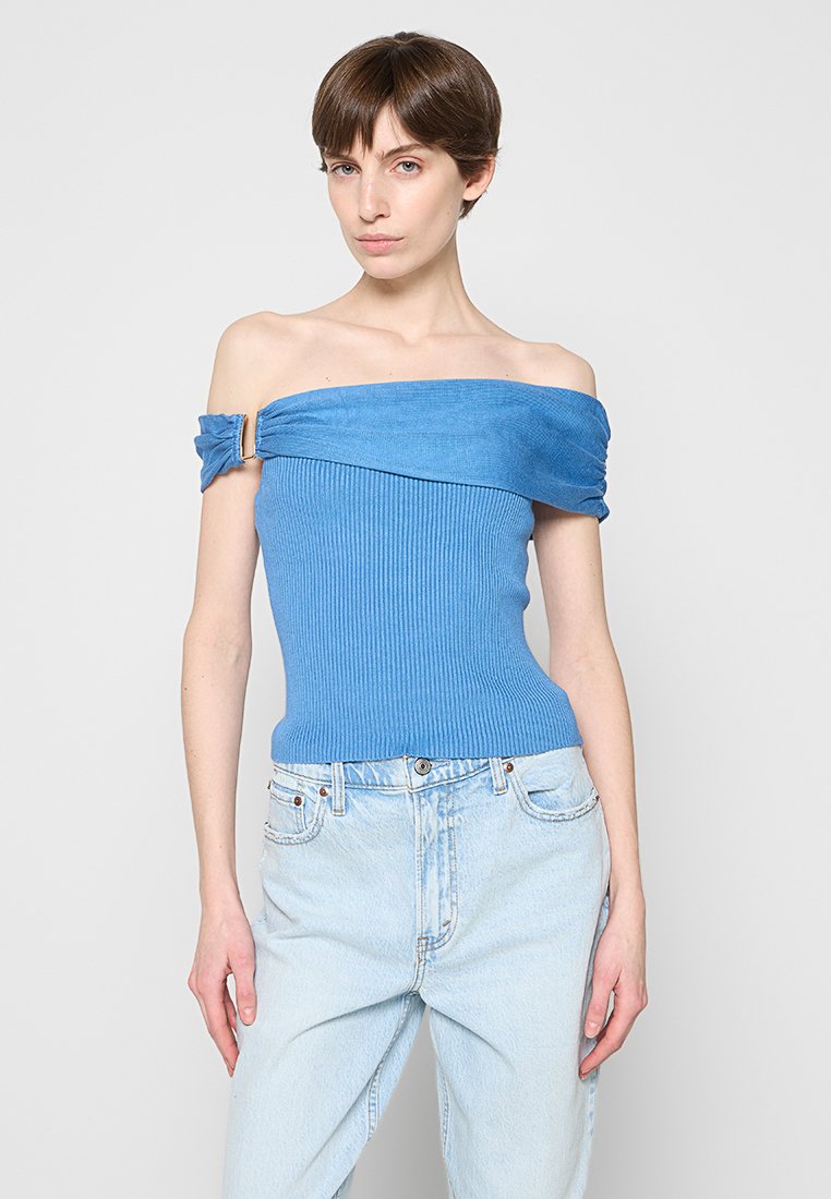 Frau mit kurzen braunen Haaren, die ein blaues, schulterfreies, geripptes Oberteil und hellblaue, hoch taillierte Jeans trägt, steht vor einem schlichten Hintergrund.