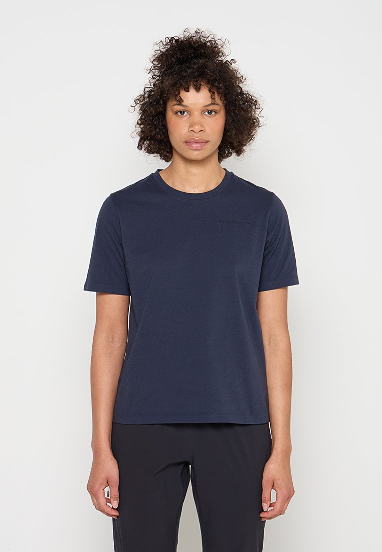Junge Person mit lockigem Haar, die ein schlichtes marineblaues T-Shirt mit kurzen Ärmeln und schwarze Hose trägt, steht vor einem hellen Hintergrund.