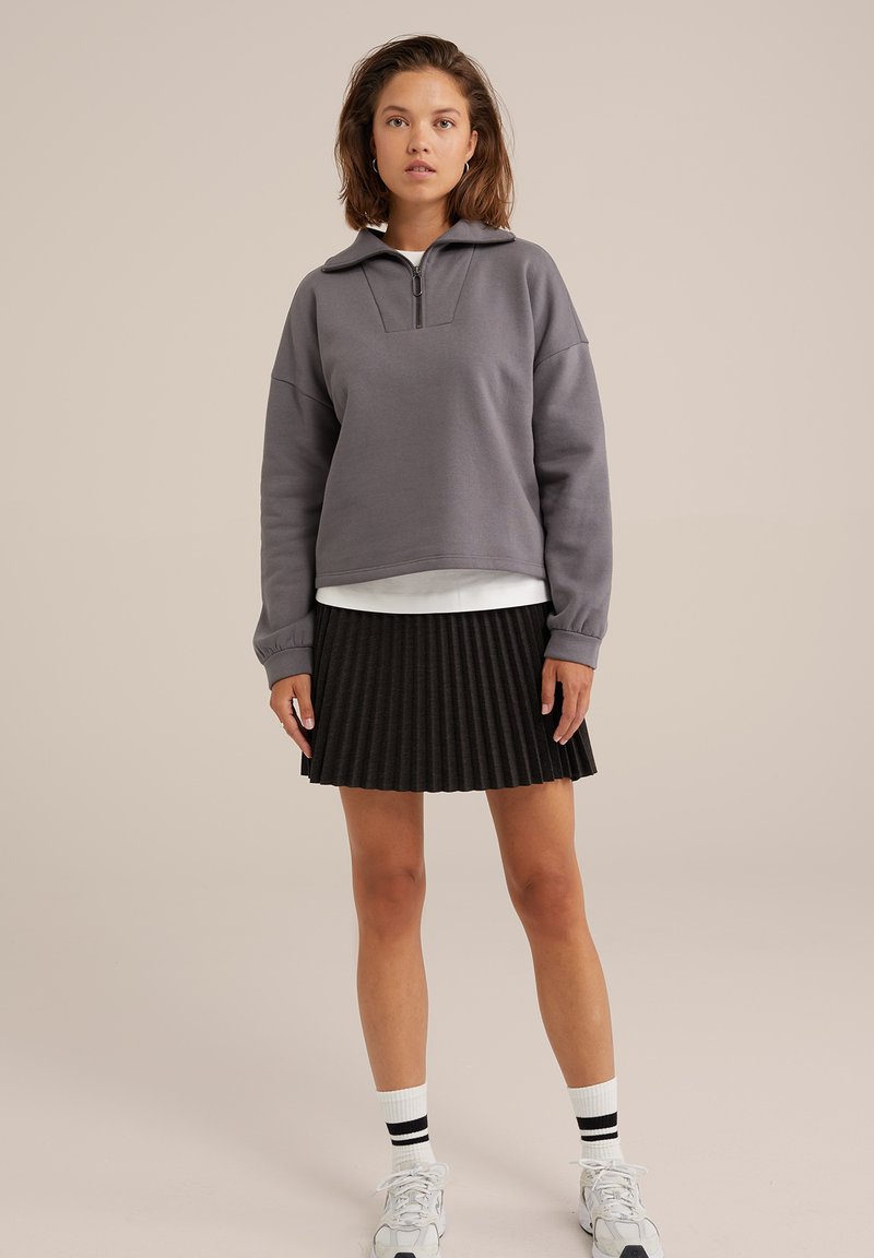 Grauer Baumwollpullover mit Reißverschlusskragen, kombiniert mit einem schwarzen plissierten Rock. Das Model trägt weiße Socken mit schwarzen Streifen und sportliche Schuhe.