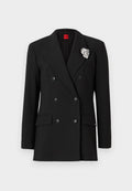 Schwarzer doppelreihiger Blazer mit großen Revers, zwei Fronttaschen und einem silbernen Broschenakzent am Revers. Glatte Stofftextur.
