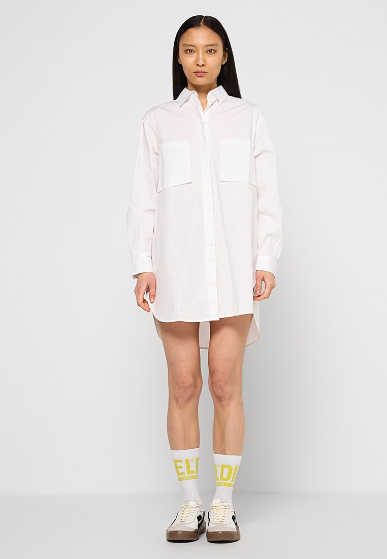 Weißes oversized Hemd mit Knopfleiste, zwei Brusttaschen und langen Ärmeln, getragen mit knöchellangen Socken und Turnschuhen. Minimalistisches Design.