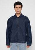 Mann trägt eine dunkelblaue Jeansjacke mit Reißverschluss über blau-weiß gestreiften Hosen und steht vor einem schlichten hellgrauen Hintergrund.