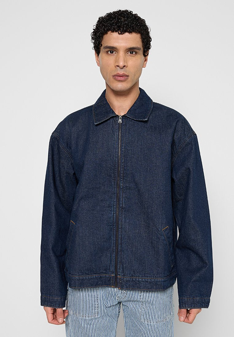 Mann trägt eine dunkelblaue Jeansjacke mit Reißverschluss über blau-weiß gestreiften Hosen und steht vor einem schlichten hellgrauen Hintergrund.