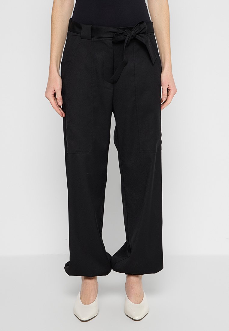 Schwarze, weit geschnittene Pants mit gebundenem Bund, ausgestattet mit zwei Fronttaschen und glatter Textur, kombiniert mit spitzen weißen Absatzschuhen.