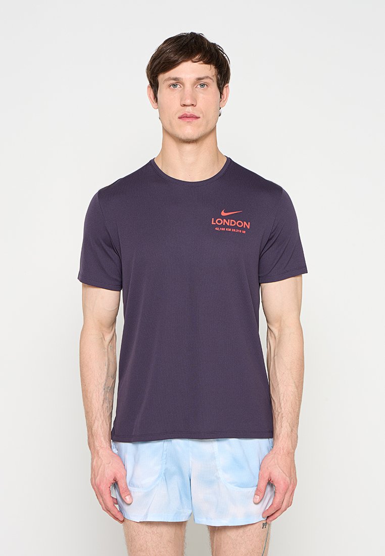 Mann trägt ein dunkelviolettes Nike London T-Shirt und hellblaue Shorts, steht vor einem schlichten weißen Hintergrund.