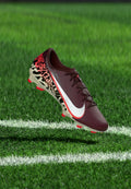 Bordeauxrote und cremefarbene Fußballschuh mit rot-schwarz gesprenkeltem Design, weißem Swoosh-Logo, schwebend über grünem Gras nahe der weißen Spielfeldlinie.