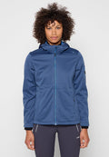 Person mit lockigem Haar, die eine blaue Outdoorjacke mit Reißverschluss und dunkle Sporthose trägt, steht vor einem einfarbigen hellen Hintergrund.