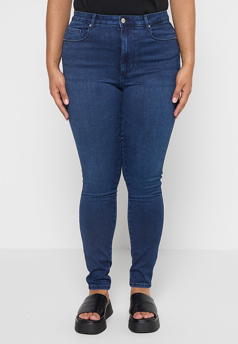 Schmale Jeans aus dunkelblauem Denim mit Taschen, einem Reißverschluss und einem Knopfverschluss. Getragen mit schwarzen Riemchensandalen.