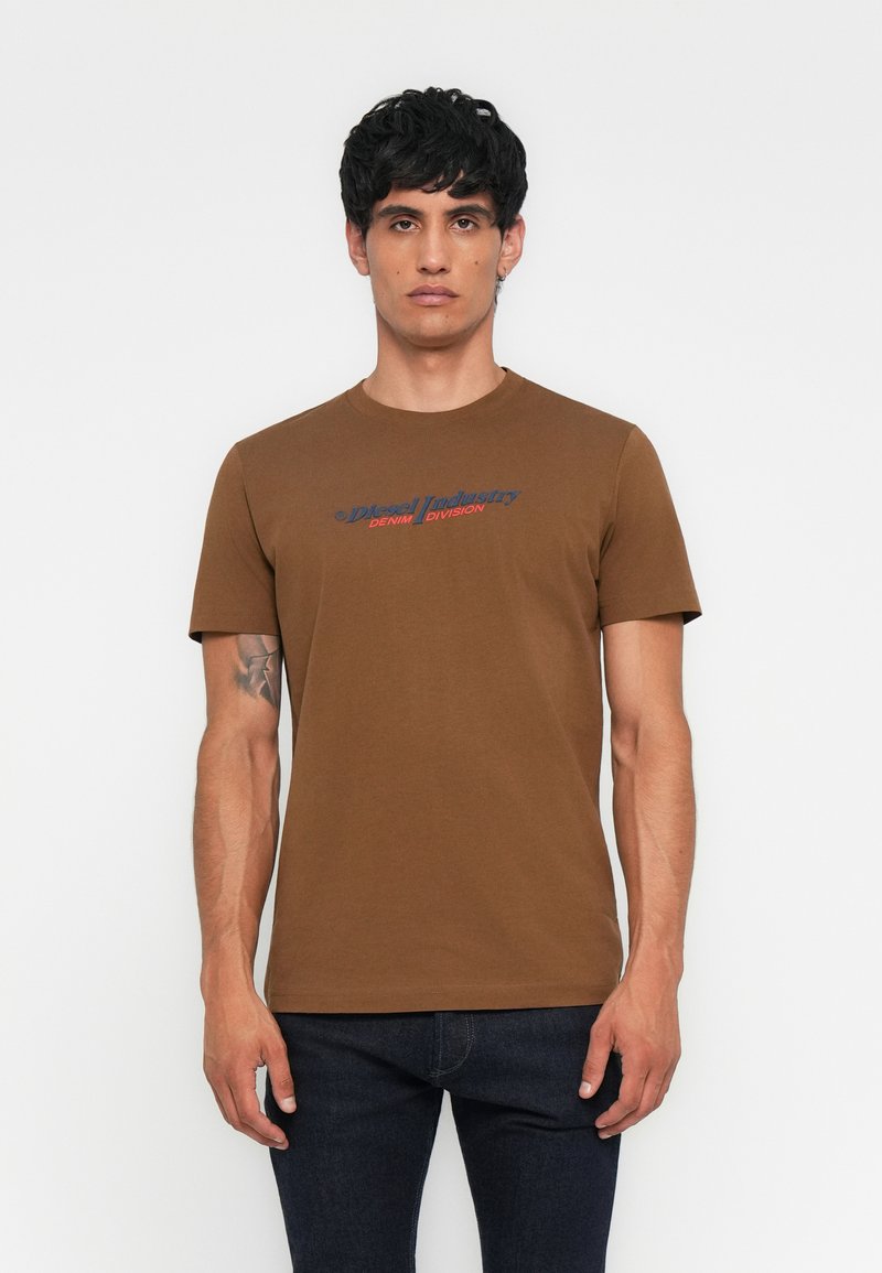 Mann mit dunklen Haaren trägt ein braunes T-Shirt von Diesel Industry Denim Division und dunkle Jeans, steht vor einem schlichten weißen Hintergrund.