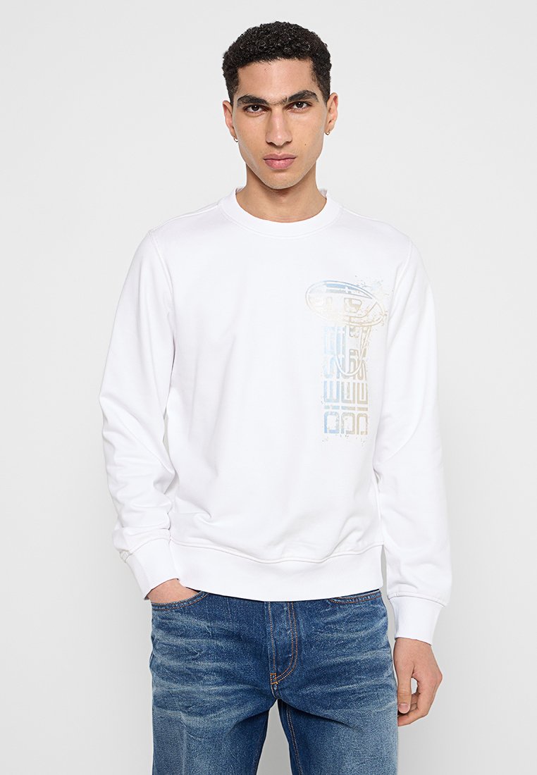 Junger Mann mit kurzen lockigen Haaren, der einen weißen Sweatshirt mit vertikal verblasstem Muster und blaue Jeans trägt, während er mit einer Hand in der Tasche steht.