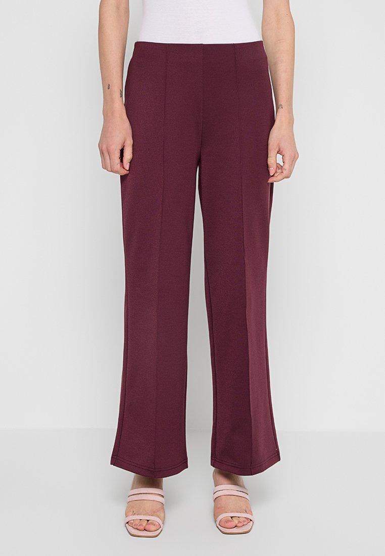 Bordeauxfarbene weit geschnittene Hose aus weichem Stoff, mit einer glatten Textur und zwei vorderen Nähten. Kombiniert mit hellrosa Riemchensandalen.