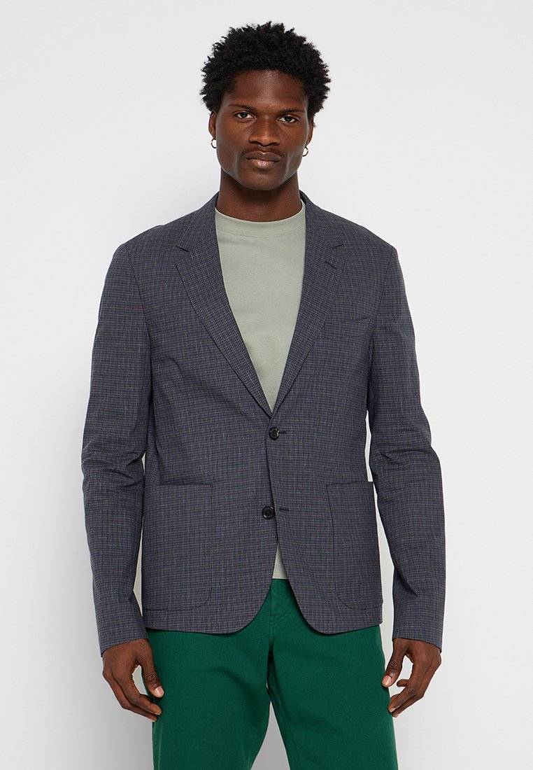 Mann in einem grauen karierten Blazer über einem hellgrünen Hemd und grünen Hosen, der vor einem schlichten weißen Hintergrund steht.