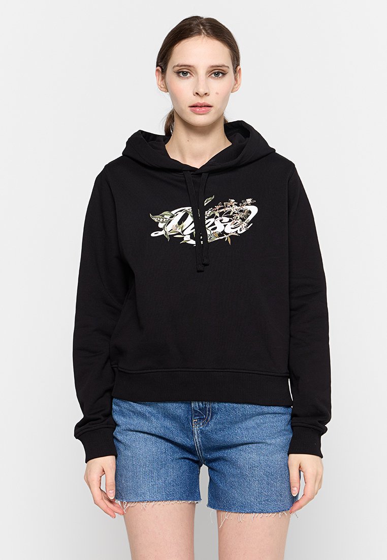 Schwarzer Kapuzenpullover mit einem grafischen Design auf der Vorderseite, das botanische Elemente zeigt. Kombiniert mit Denim-Shorts, die einen lässigen Stil präsentieren.