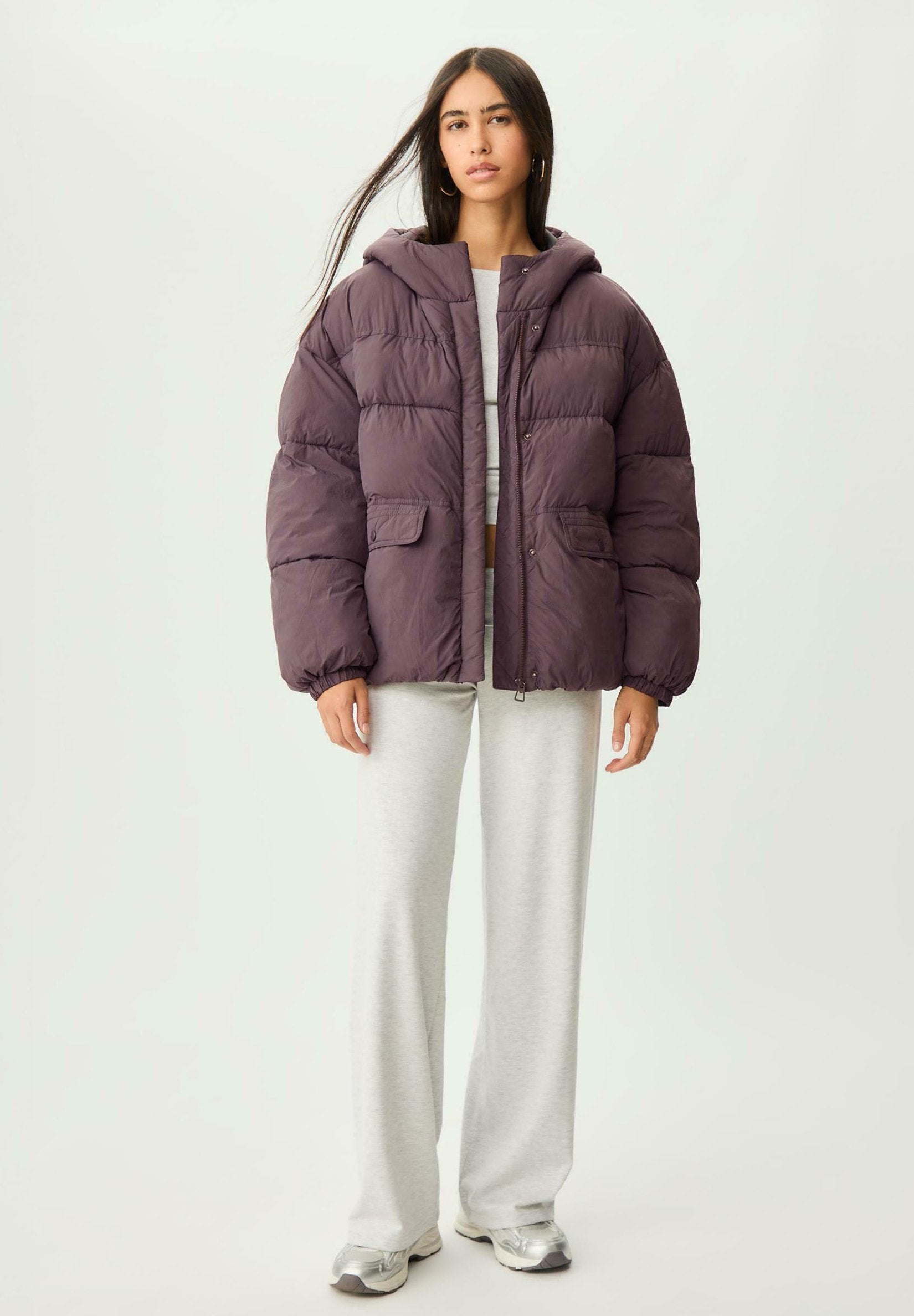 Dunkelviolette Pufferjacke mit Kapuze, zwei vorderen Taschen und glatter Textur, kombiniert mit hellgrauen Sweatpants und metallischen Sneakers.