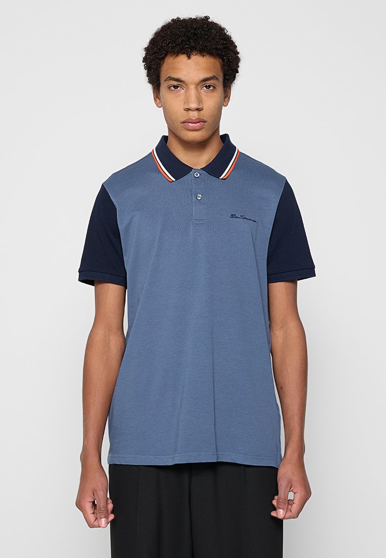 Polohemd in Blau mit einem einfarbigen Marinekragen, der rot-weiße Streifen hat. Weicher Stoff, kurze Ärmel und ein kleines Logo auf der Brust.
